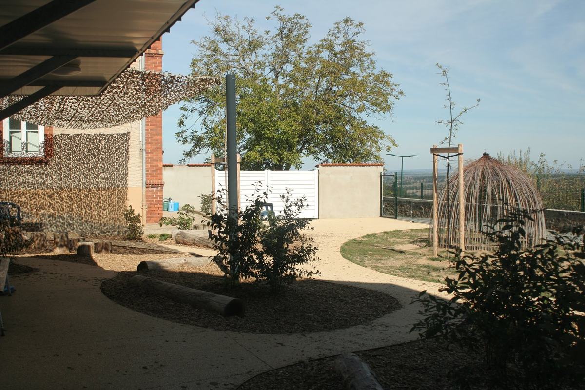 Abri en forme de dôme tressé en osier dans la cour de récréation de l'école, entouré d'arbres et de rondins de bois.