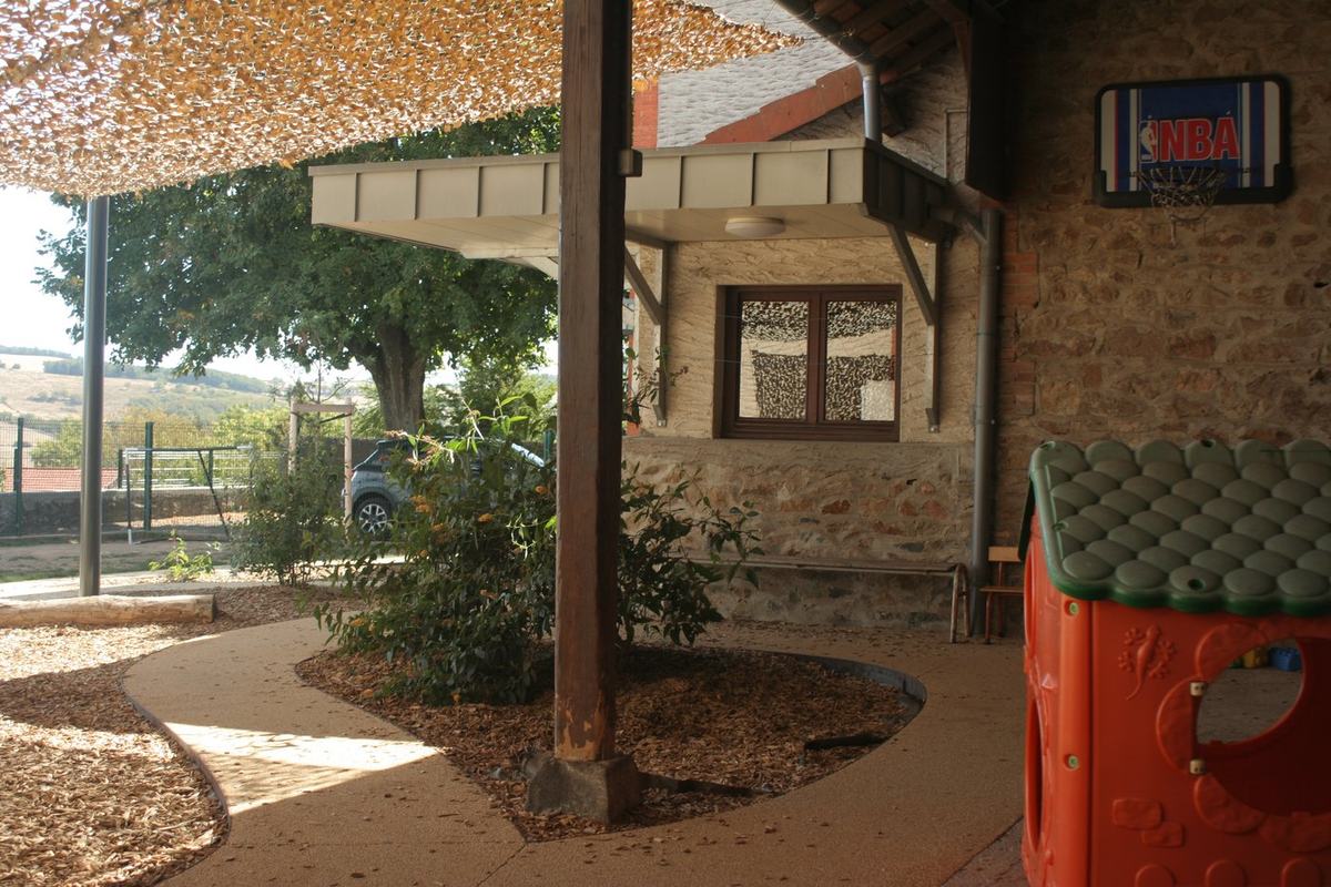 Préau de l'école avec panier de basket NBA fixé au mur en pierre et structure de jeux pour enfants.