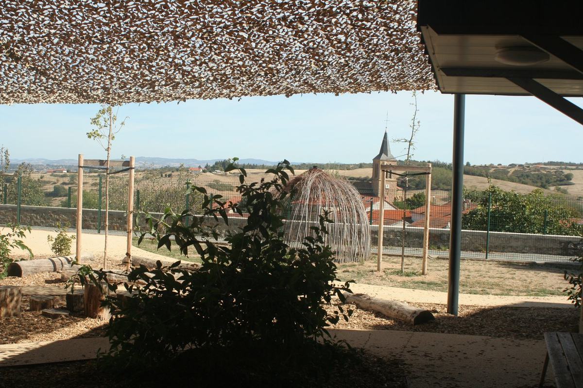 Cour de récréation de l'école aménagée avec une voile d'ombrage, des rondins de bois et un abri en osier tressé, avec vue sur le clocher et les toits du village.