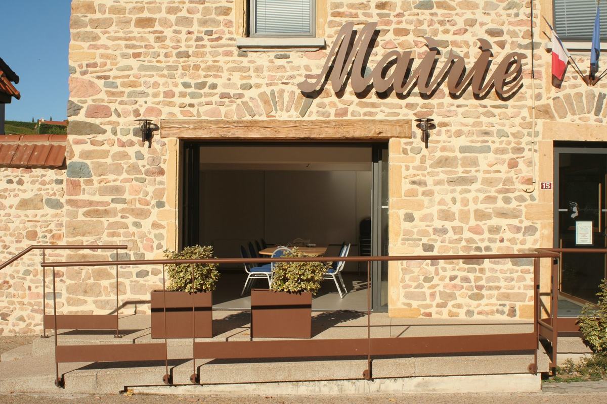 Façade en pierre de la mairie de Vendranges avec l'enseigne « Mairie », les drapeaux français et européen, et la salle Louis-Ranvier visible en rez-de-chaussée, baie vitrée ouverte sur la terrasse.