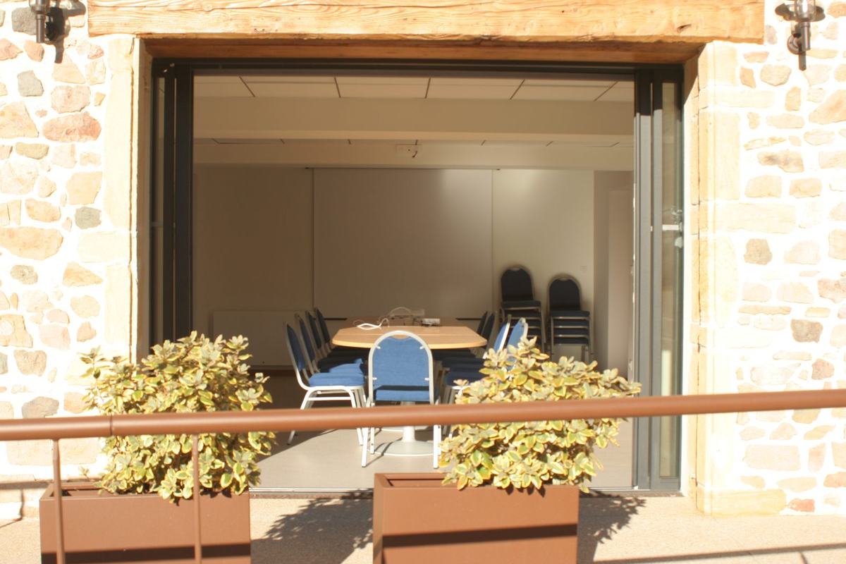 Vue depuis la terrasse de la mairie de Vendranges à travers la baie vitrée ouverte de la salle Louis-Ranvier, avec sa table de réunion et ses chaises.